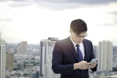 Takım elbiseli genç Asyalı işadamı gökdelen manzaralı çatıda akıllı telefon kullanıyor.