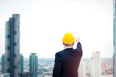 Güvenlik kaskı takmış Asyalı genç işadamı gökyüzünü gökdelen şehriyle göstererek gelecek konseptine gidiyor. 