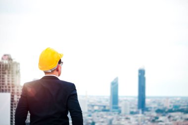 Güvenlik kaskı takmış iş adamının sırtında gökyüzüne gökdelen şehriyle bakıyor ve geleceğe doğru yol alıyor. 