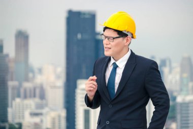 Güvenlik kaskı takmış, çatıda gökdelen manzaralı şarkı söyleyip dans eden Asyalı genç işadamı.