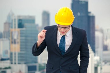 Güvenlik kaskı takmış, çatıda gökdelen manzaralı şarkı söyleyip dans eden Asyalı genç işadamı.