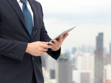 Takım elbiseli genç Asyalı işadamı çatıda dijital tablet kullanıyor ve gökdelen şehir manzaralı.