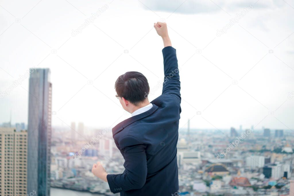Cheerful and happy achievement successful young Asian businessman in suit raising his arm up to celebrate success goal on the rooftop with skyscraper city view