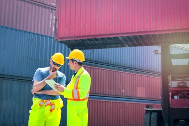 Güvenlik giysileri ve miğfer giyen iki işçi arka plan olarak lojistik kargo konteynırları deposundaki forklift kamyon konteynır kutusuyla ilgili bir şeyi tartışıyor.