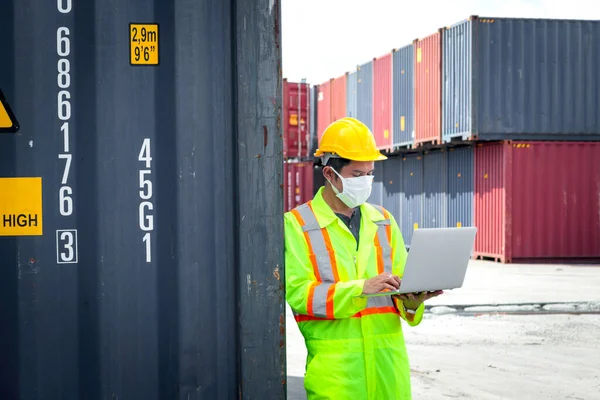 Worker with protection face mask wearing safety clothing and helmet standing at logistic shipping yard and using laptop computer near cargo containers box