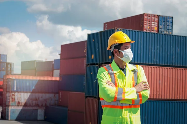 Worker with protection face mask wearing safety clothing and helmet standing with crossed his arms at logistic cargo containers shipping yard
