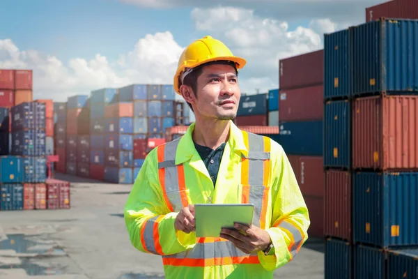 Worker with protection face mask wearing safety clothing and helmet standing using digital tablet at logistic cargo containers shipping yard