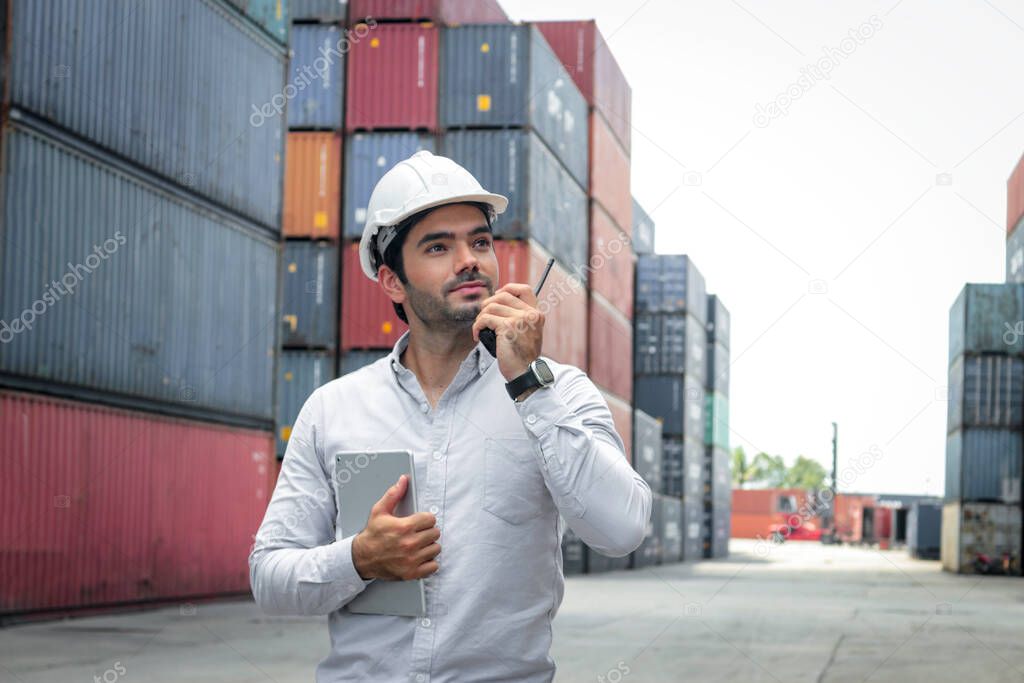 Handsome businessman wearing safety helmet,  using walkie talkie and hold digital table during inspection at logistic shipping cargo containers yard