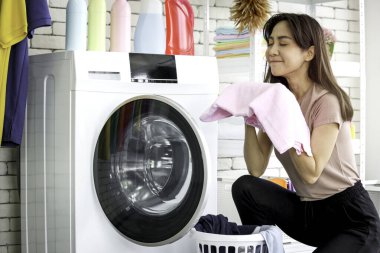 Mutlu gülümseyen Asyalı güzel bir kadın çamaşır odasında çamaşır makinesiyle çamaşır yıkıyor ev hanımı ev işi yapıyor ve evde çamaşır yıkıyor.