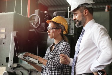 Genç ve güzel endüstriyel mühendis kadın miğfer ve güvenlik gözlüğü takıyor iş adamıyla tartışıyor, dizüstü bilgisayar kullanıyor, imalat fabrikasında çalışıyor, patron ve sekreter.  