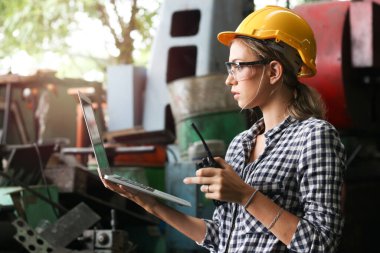 Miğfer takan sanayi mühendisi ve güvenli gözlük takan, elinde telsiz olan, imalat fabrikasında dizüstü bilgisayar kullanan, endüstride çalışan genç ve güzel bir kadın. 