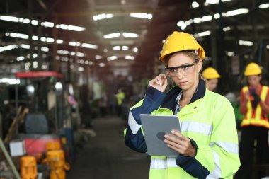 Genç, güzel, endüstriyel mühendis kadın, kaskını ve koruyucu giysilerini giyiyor. Fabrika imalatı için dijital tablet taşıyor. İş sektörü konsepti.