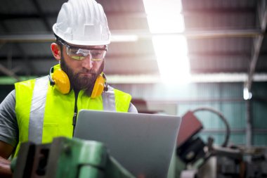 Kasklı ve güvenli gözlük takan endüstri mühendisi, fabrika fabrikasındaki işletim makineleri için dizüstü bilgisayar tutuyor, endüstri konseptinde makine ile çalışıyor. 