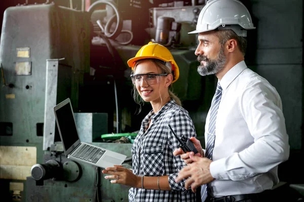 Young beautiful woman industrial engineer worker wearing helmet and safety glasses discussing with businessman, using laptop computer, people work at manufacturing industry factory, boss & secretary