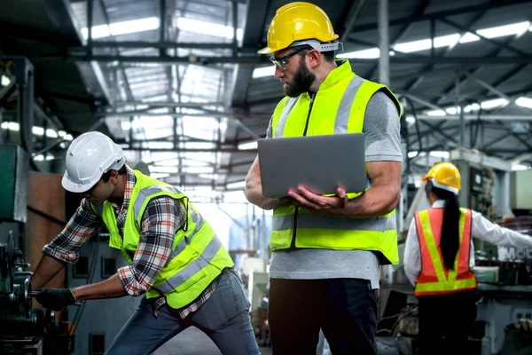 Industrial engineer wearing helmet and safe glasses, holding laptop computer for operating machinery at manufacturing plant factory, worker team working together in industry concept