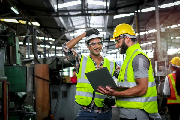 Industrial engineer wearing helmet and safe glasses, holding laptop computer for operating machinery at manufacturing plant factory, worker team working together in industry concept