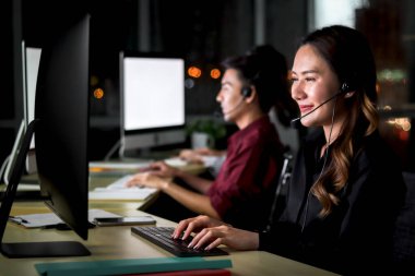 Mutlu, gülümseyen, kulaklıklı, çağrı merkezinde çalışan, takım arkadaşlarıyla birlikte çalışan, elleri boş, mutlu iş yeri konsepti olan, genç, güzel, Asyalı bir kadın.