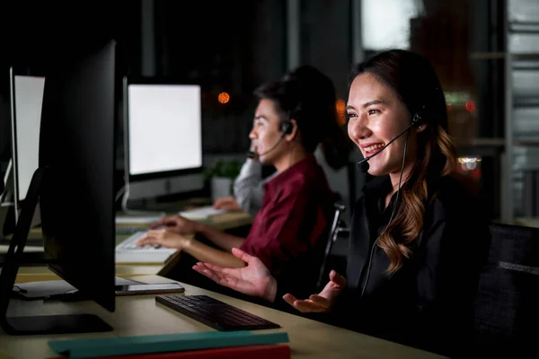 Happy smiling young beautiful Asian woman with headphones working at call center service desk consultant with her teammates at night, ready to take with customer on hands-free phone, happy workplace concept