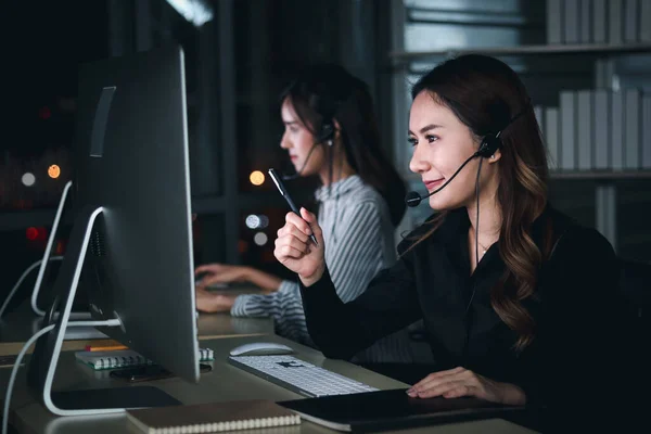 Happy smiling young beautiful Asian woman with headphones working at call center service desk consultant with her teammates at night, pointing at computer screen, taking with customer on hands-free phone, happy workplace concept