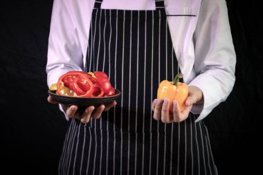 Şef önlük giyiyor, siyah arka planda taze dolmalık biber tutuyor, sağlıklı yiyecek kavramı var.