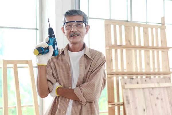 Portrait of senior man carpenter holding  the hand drill, standing in carpentry woodwork  workshop, old craftsman enjoy his DIY hobby