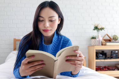 Beyaz yatak odasında rahat bir yatakta uzanırken Asyalı bir kadının kitap okuması. Evde roman okuyan mutlu genç bir kız. Güzel dişiler boş zamanlarında eğlenmeyi severler..