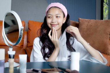 Beautiful female teen in bathrobe doing treatment face with skincare hydrating mask at home. Happy teenage Asian girl look in mirror, apply cosmetic on face. Beauty healthcare treatment cosmetics.