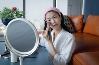 Beautiful female teen in bathrobe applying makeup cosmetic on her face at home. Happy teenage Asian girl brushing her cheek while looks in the mirror. Beauty healthcare treatment and makeup cosmetics.
