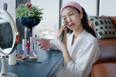 Beautiful female teen in bathrobe applying makeup cosmetic on her face at home. Happy teenage Asian girl brushing her cheek while looks in the mirror. Beauty healthcare treatment and makeup cosmetics.