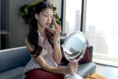 Beautiful female teen applying makeup cosmetic on her face at home. Happy teenage Asian girl brushing her cheek while looks in the mirror. Beauty healthcare treatment and makeup cosmetics.