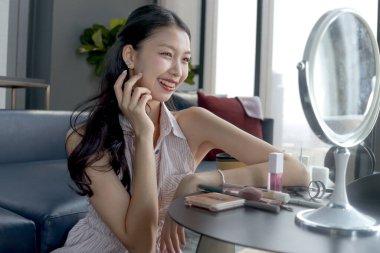 Female teenager looks at her beautiful appearance in mirror after applying makeup cosmetic on face at home. Happy teen Asian girl checking her appearance after finishing makeup. Beauty makeup cosmetic