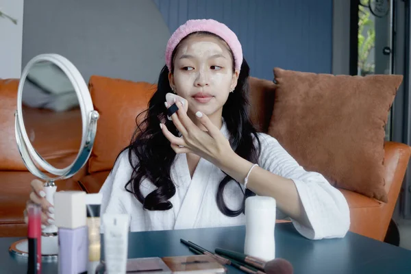 Beautiful female teen in bathrobe holding mirror and applying cosmetic on her face at home. Happy teenage Asian girl applies makeup on face while looks in mirror. Beauty healthcare treatment cosmetics