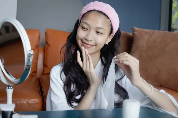Beautiful female teen in bathrobe doing treatment face with skincare hydrating mask at home. Happy teenage Asian girl look in mirror, apply cosmetic on face. Beauty healthcare treatment cosmetics.