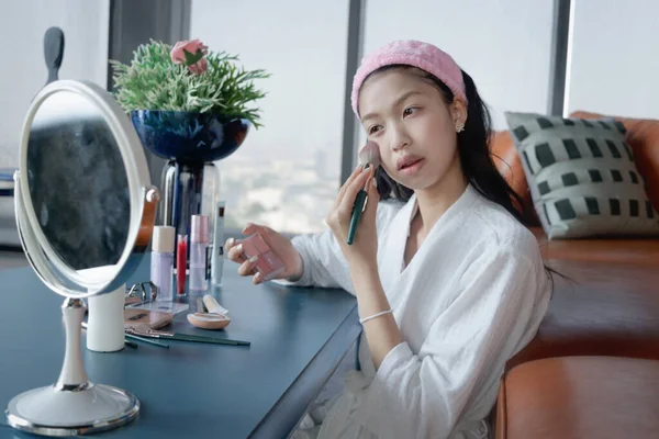 Beautiful female teen in bathrobe applying makeup cosmetic on her face at home. Happy teenage Asian girl brushing her cheek while looks in the mirror. Beauty healthcare treatment and makeup cosmetics.