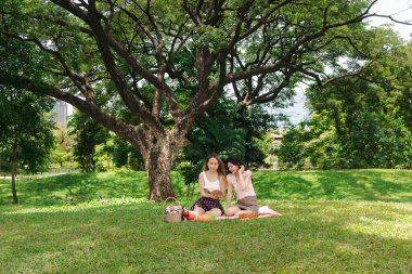 Mutlu gülümseyen iki güzel Asyalı kadın yeşil parkta piknik yapıyor. Yakın arkadaş kızlar açık hava pikniğinde kitap okuyor. Rahatlayan insanlar doğal ortamlarda birlikte vakit geçirirler..