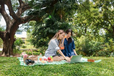 İki güzel Asyalı kadın yeşil parkta piknik yapıyor. Yakın arkadaş kızları rahatlatmak portakal suyu içmek ve dışarıda piknik yaparken dizüstü bilgisayar kullanmak. İnsanlar doğada birlikte vakit geçirirler..