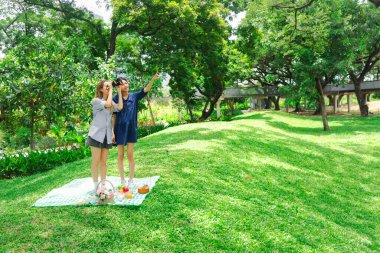 Mutlu iki güzel Asyalı kadın doğa yeşili parkta piknik yapıyor. Yakın arkadaş kızlar açık hava pikniğinde dürbünle kuş gözlemlemeye bakarlar. İnsanlar doğal ortamlarda birlikte vakit geçirirler.