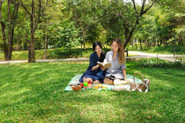 Mutlu gülümseyen iki güzel Asyalı kadın yeşil parkta piknik yapıyor. Yakın arkadaş kızlar açık hava pikniğinde kitap okuyor. Rahatlayan insanlar doğal ortamlarda birlikte vakit geçirirler..