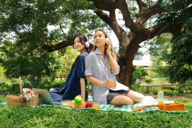 Mutlu iki güzel Asyalı kadın yeşil parkta piknik yapıyor. Kulaklıklı yakın arkadaşlar piknik sırasında dışarıda müzik dinlemek için cep telefonu kullanırlar. Rahatlayan insanlar doğada birlikte vakit geçirirler.