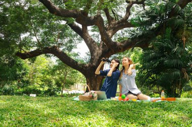 Mutlu iki güzel Asyalı kadın doğa yeşili parkta piknik yapıyor. Yakın arkadaş kızlar açık hava pikniğinde dürbünle kuş gözlemlemeye bakarlar. İnsanlar doğal ortamlarda birlikte vakit geçirirler.