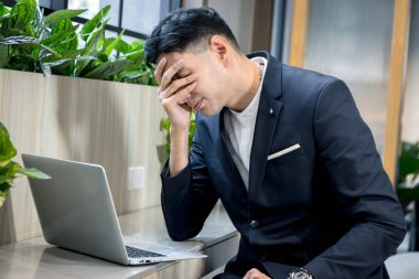 Takım elbiseli genç işadamı dizüstü bilgisayarda çalışırken sıkılmış ve stresli hissediyor. Başı ağrıyan iş adamları, yeşil modern ofiste çalışırken kafa kafaya veriyorlar..