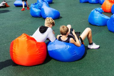 Ellerinde akıllı telefon olan gençler dinlenmek için uzayda fasulye çuvallarının üzerine otururlar. Gençler açık havada yumuşak koltuklarda rahatlar..