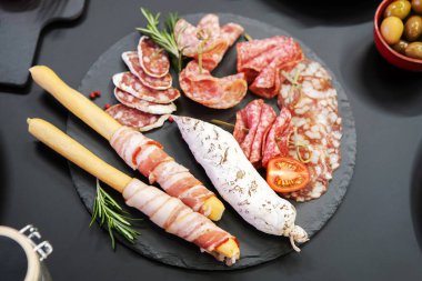 Domates ve otlarla süslenmiş siyah bir tabakta sosis lezzetleri. Et ürünleri tatmak.