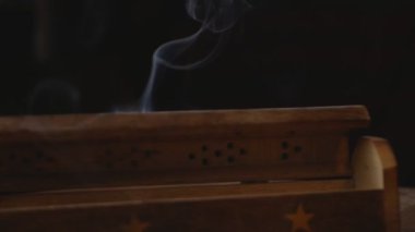 Tütsü (tütsü çubukları), yıldızlı eşsiz bir ahşap kutudan tüttürür. Duman deliklerden çıkıyor. Kutu el yapımı bir masanın üzerinde. Saat yönünde dönüyor. Dumana odaklan. Siyah arka plan. Döngü.