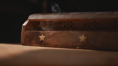 Eşsiz bir ahşap tütsüden çıkan duman tabakaları odağını kaybediyor. Duman çekmecelerden boşluklarla çıkıyor. El yapımı bir masada kutu. Kutu ışığa gelir ve karanlığa gider. Dumana odaklan. Siyah arkaplan.