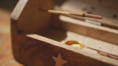 Tütsü çubukları, üzerinde yıldız olan eşsiz bir tahta kutudan tütsülenir. Yarım dönüş. Tütsü odağını kaybeder. Yakın plan. Kutudaki yıldızlar odaklanmış..