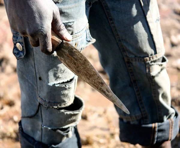 A makeshift primitive knife in the hand of a dark-skinned man. A simple carving knife in the right hand of a village African against a backdrop of torn jeans.