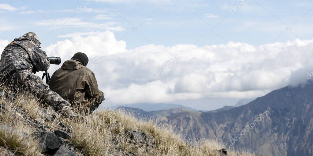 Hunters watch the animals on the opposite mountain slope with a telescope. Two men in camouflage disguise themselves in the mountains with optical devices.