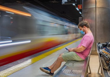 Mallorca public transport, Young man waiting for subway sitting on a bench alone with mask and safety distance due to covid-19 crisis using the phone while the subway passes in front of him