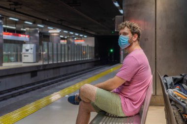 Mallorca public transport, Young man waiting for the subway sitting on a bench alone with a mask and safety distance due to the covid crisis 19, man with a red shirt and shorts looking at the platform